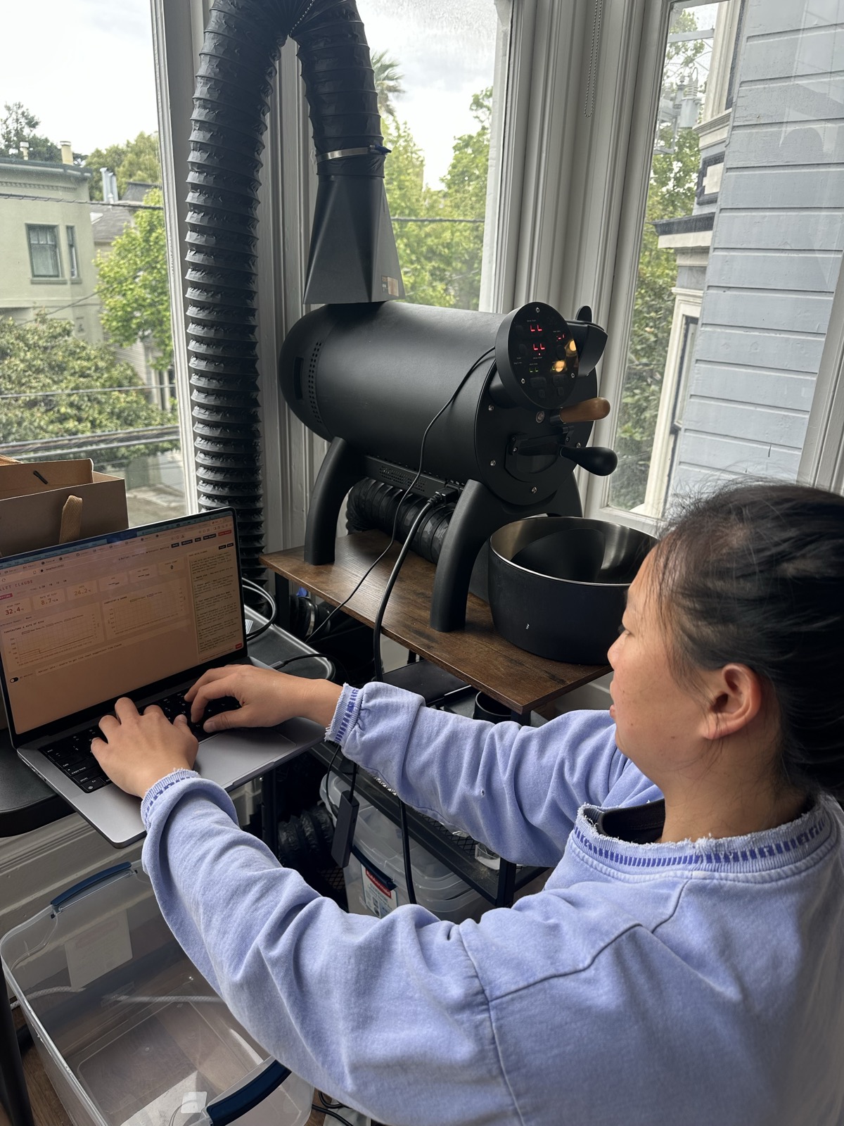 Me at a laptop next to the Aillio Bullet R1 black drum roaster, with exhaust ducting going up out a bay window.