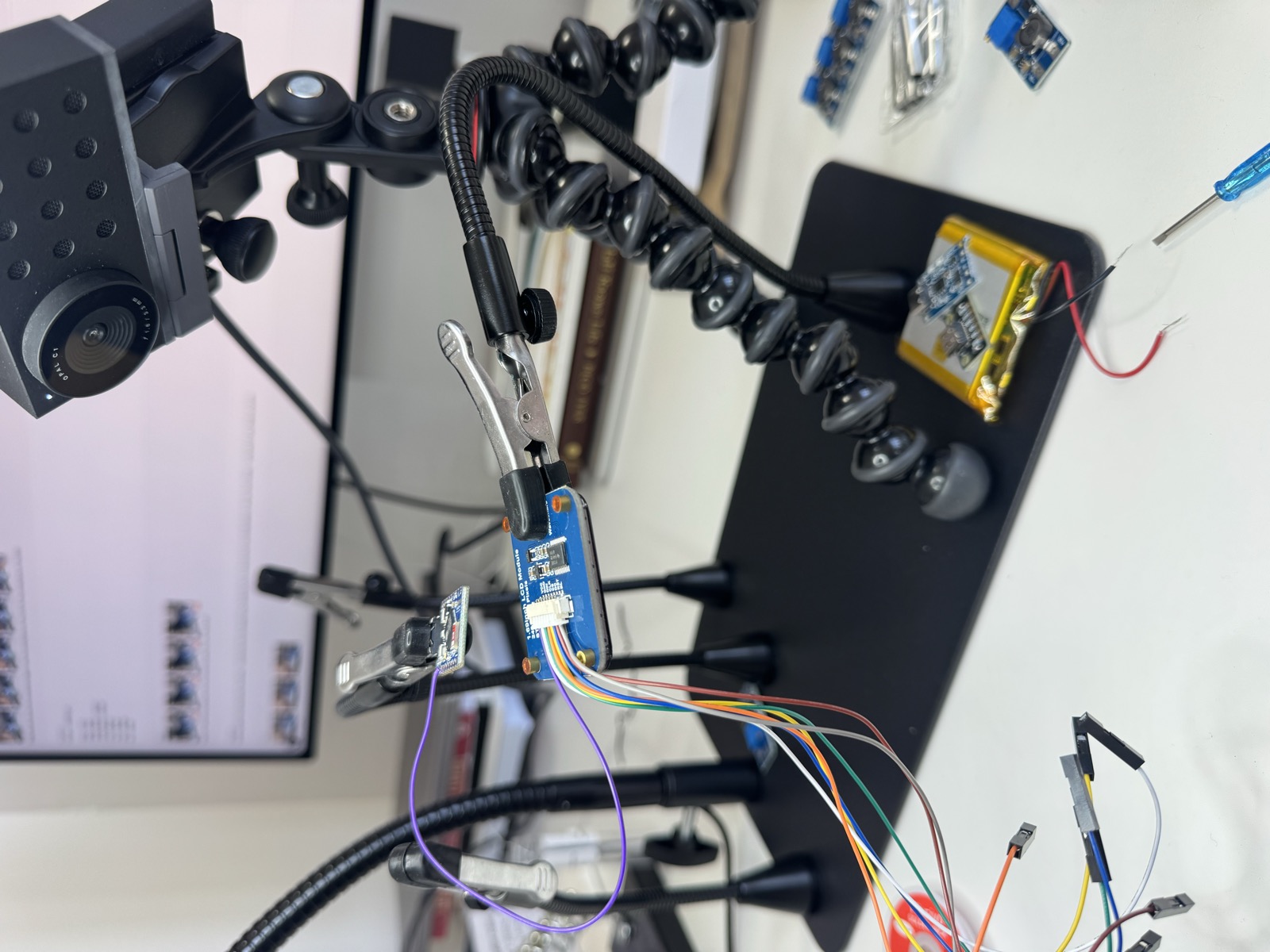 Hardware build setup — magnifying lamp, alligator-clip stands, breadboard with sensors wired up, monitor in background showing the webcam build tool.